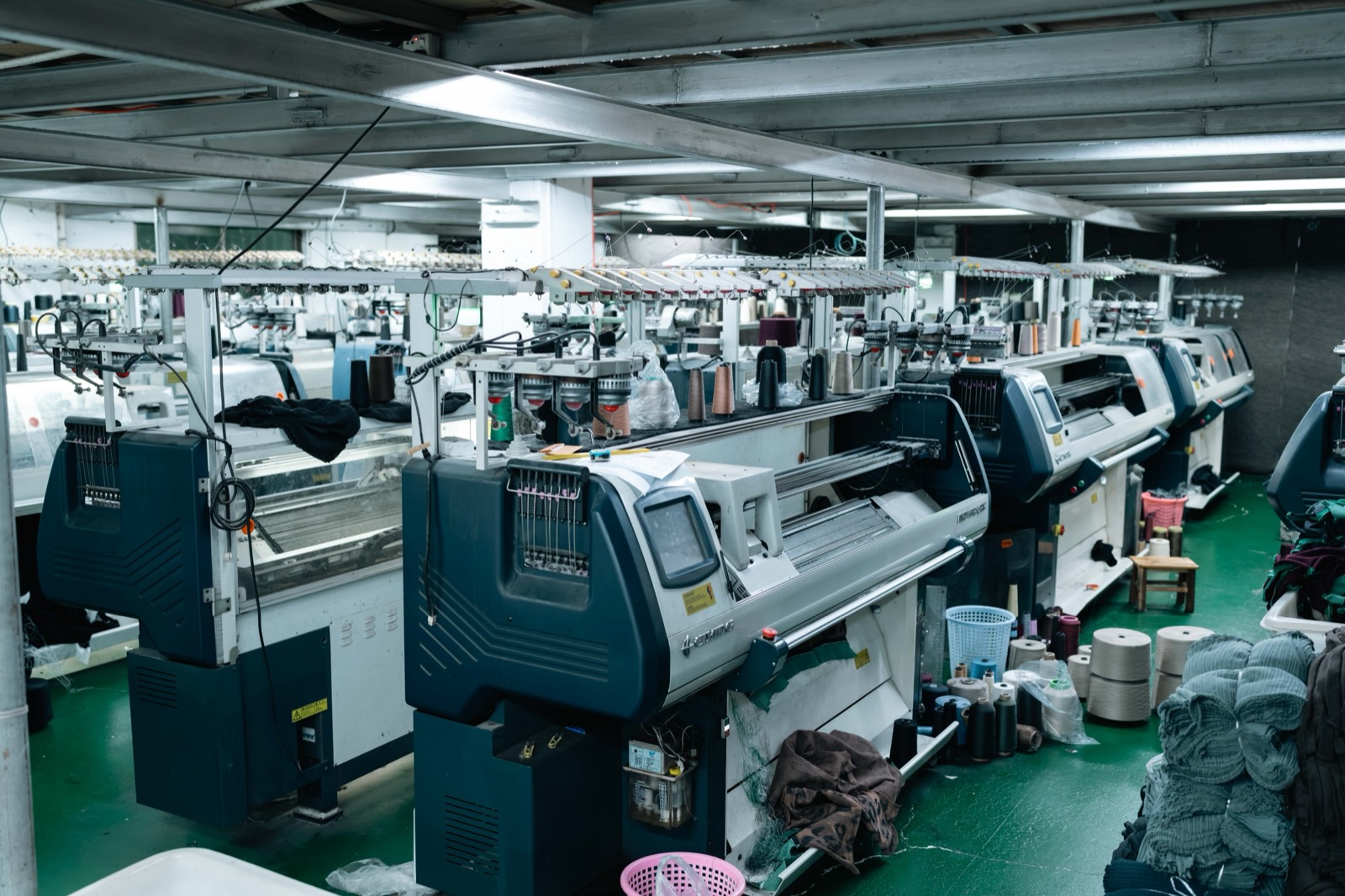 Wide view down an aisle of flat knitting machines with knitted panels collecting below